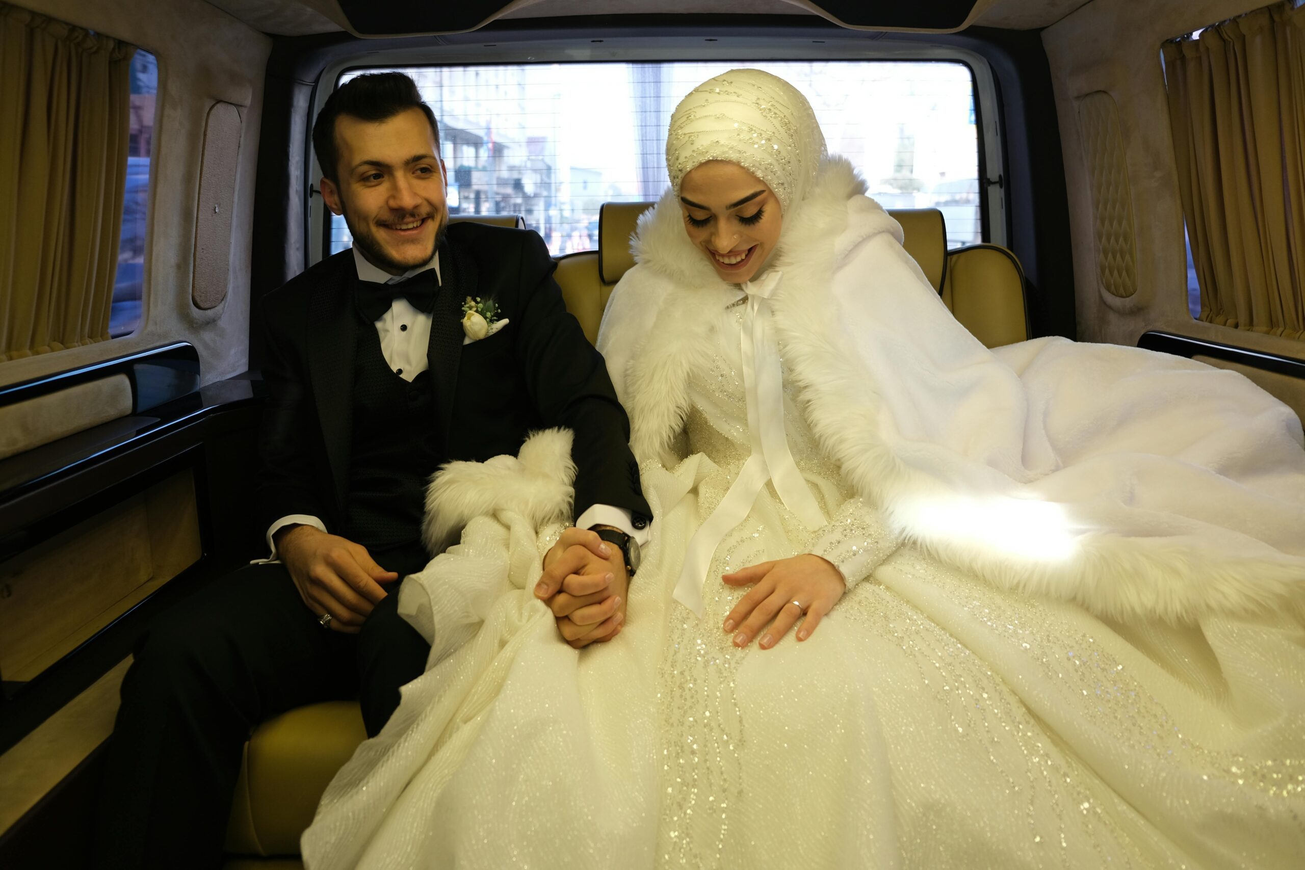 Happy newlywed couple holding hands inside a car, dressed in elegant wedding attire.
